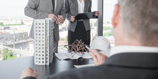 Business presentation of architects for investors, discussing new construction shapes, model of modern multi storey residential building house, blueprints and hardhat on table