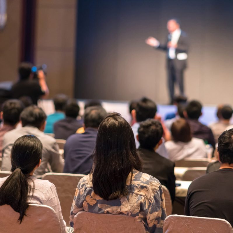 Rear side of Audiences sitting and listening the speackers on the stage in low light conference hall, event and seminar concept